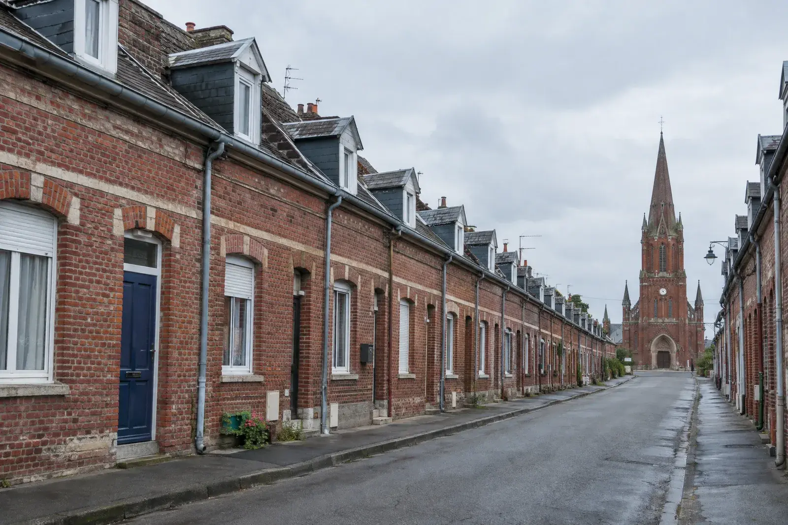 Rue de maisons ouvrières mitoyennes en brique rouge avec lucarnes et gouttières en zinc, clocher néo-gothique en brique au fond