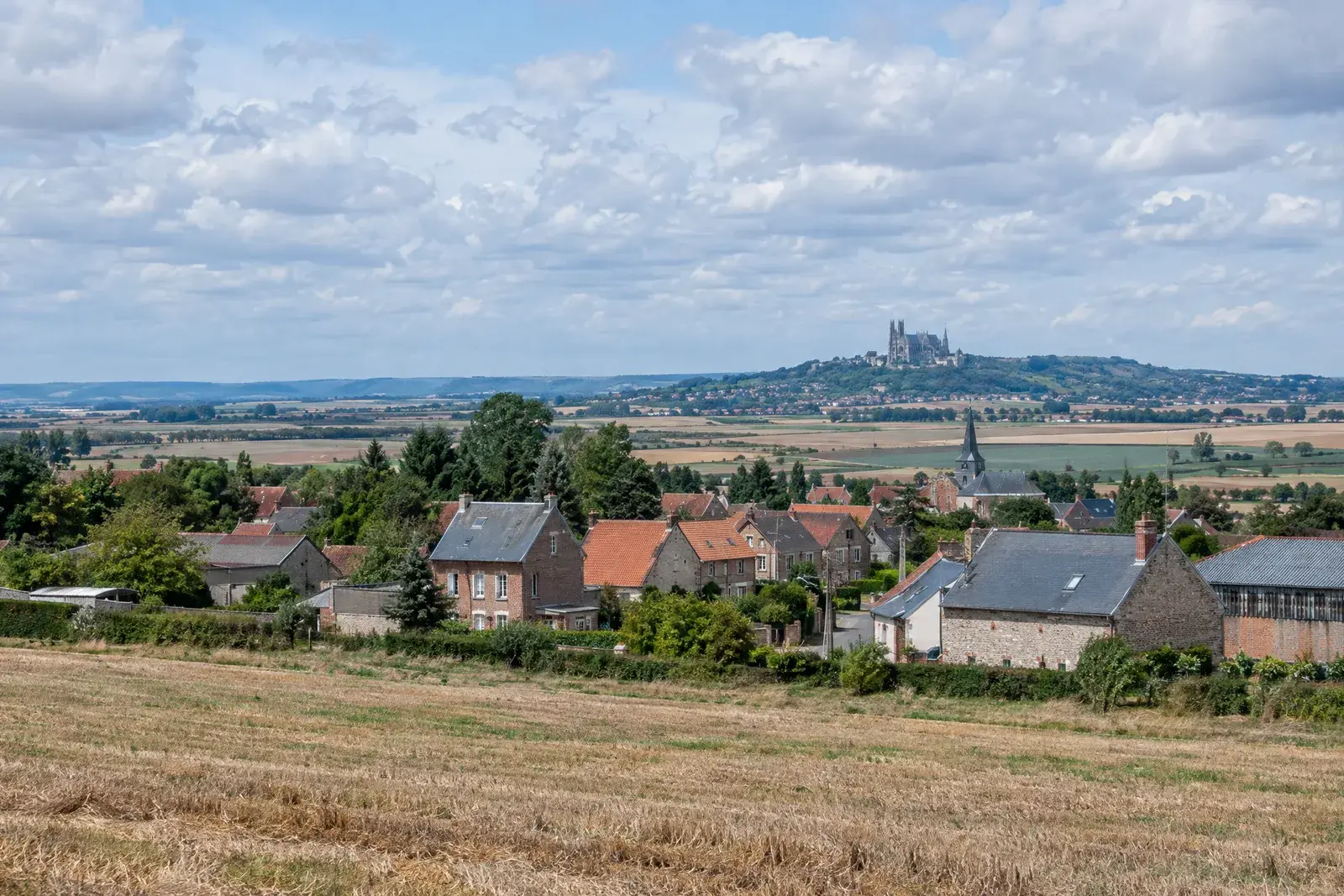 Maisons d’un village de plaine entourées de champs, une butte couronnée d’une cathédrale à l’horizon