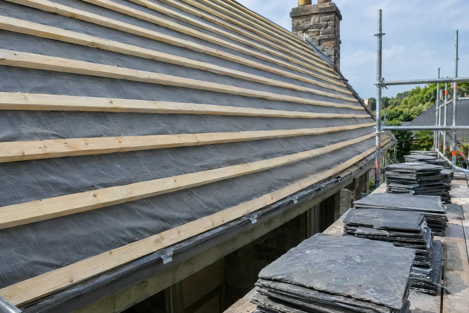 Toiture en cours de réfection : liteaux neufs cloués sur un écran de sous-toiture, ardoises neuves en piles sur l’échafaudage