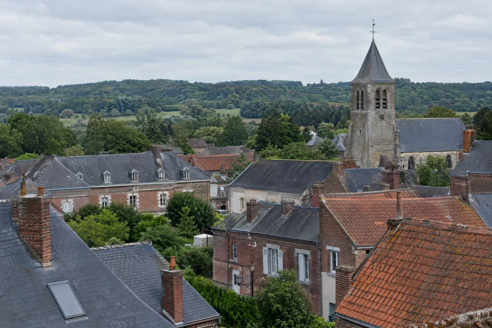 Toits de brique, d’ardoise et de tuile d’une petite ville de Thiérache, un clocher et des collines boisées à l’horizon